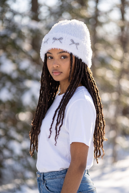 Femme portant un bonnet blanc Vélya en fausse fourrure douce avec trois nœuds décoratifs perlés dans un décor enneigé